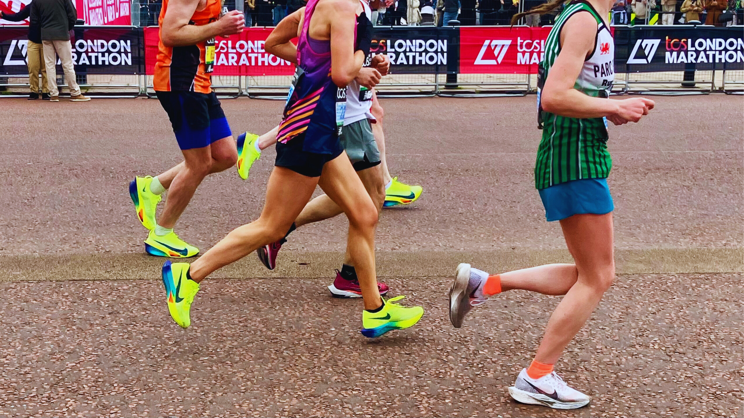 The bottom half of five runners running on tarmac. Barriers with TCS London Marathon written on them can be seen in the background, along with the legs of two people stood by the banners with their backs to the runners. 