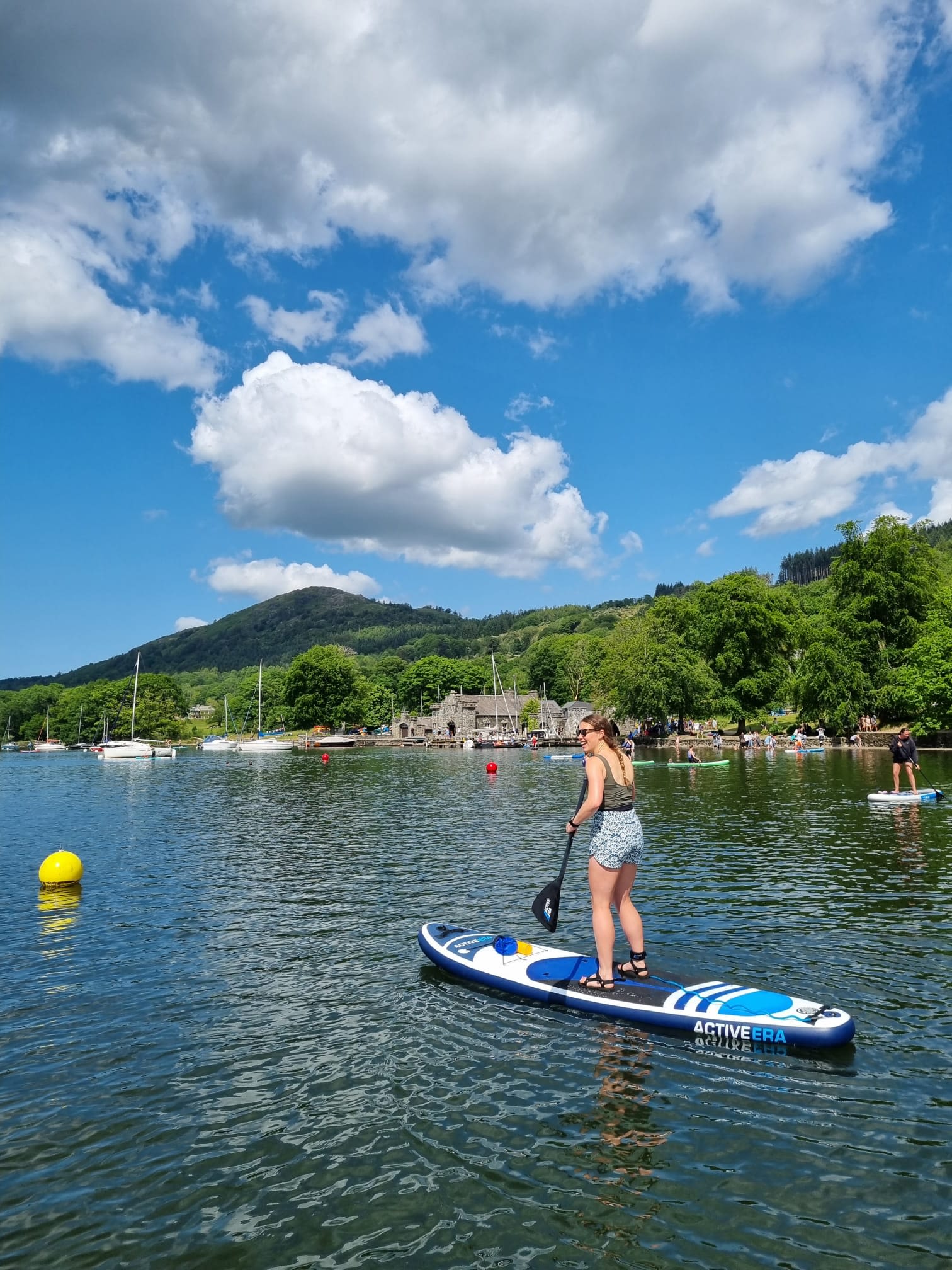 Elise paddle boarding in the Lake District with a backdrop of green hills and mountains and a blue sky 
