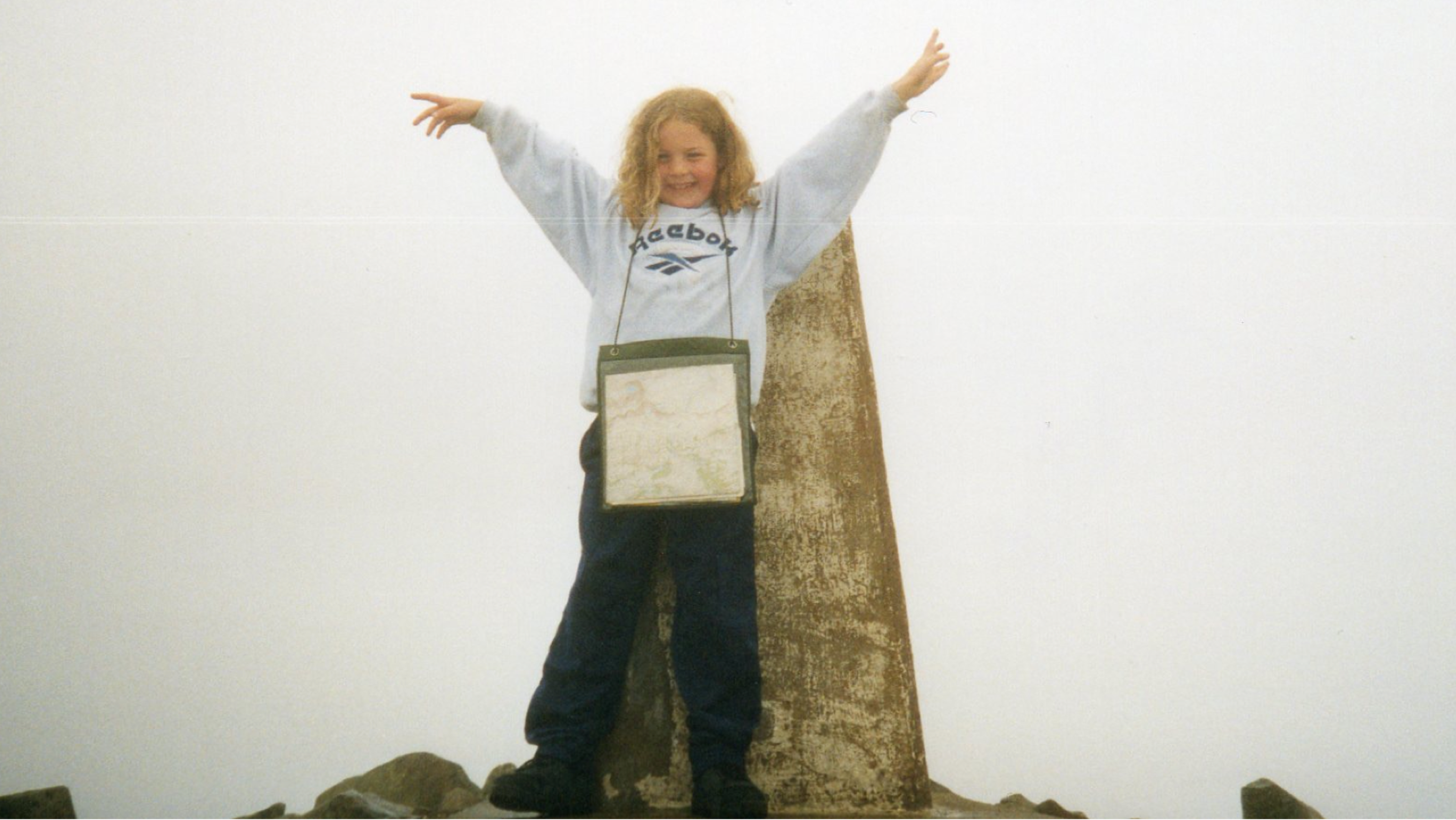 Elise age 6, wearing a grey Reebok sweatshirt, standing at the top of Cadair Idris in Wales with a map around her neck and hands in the air