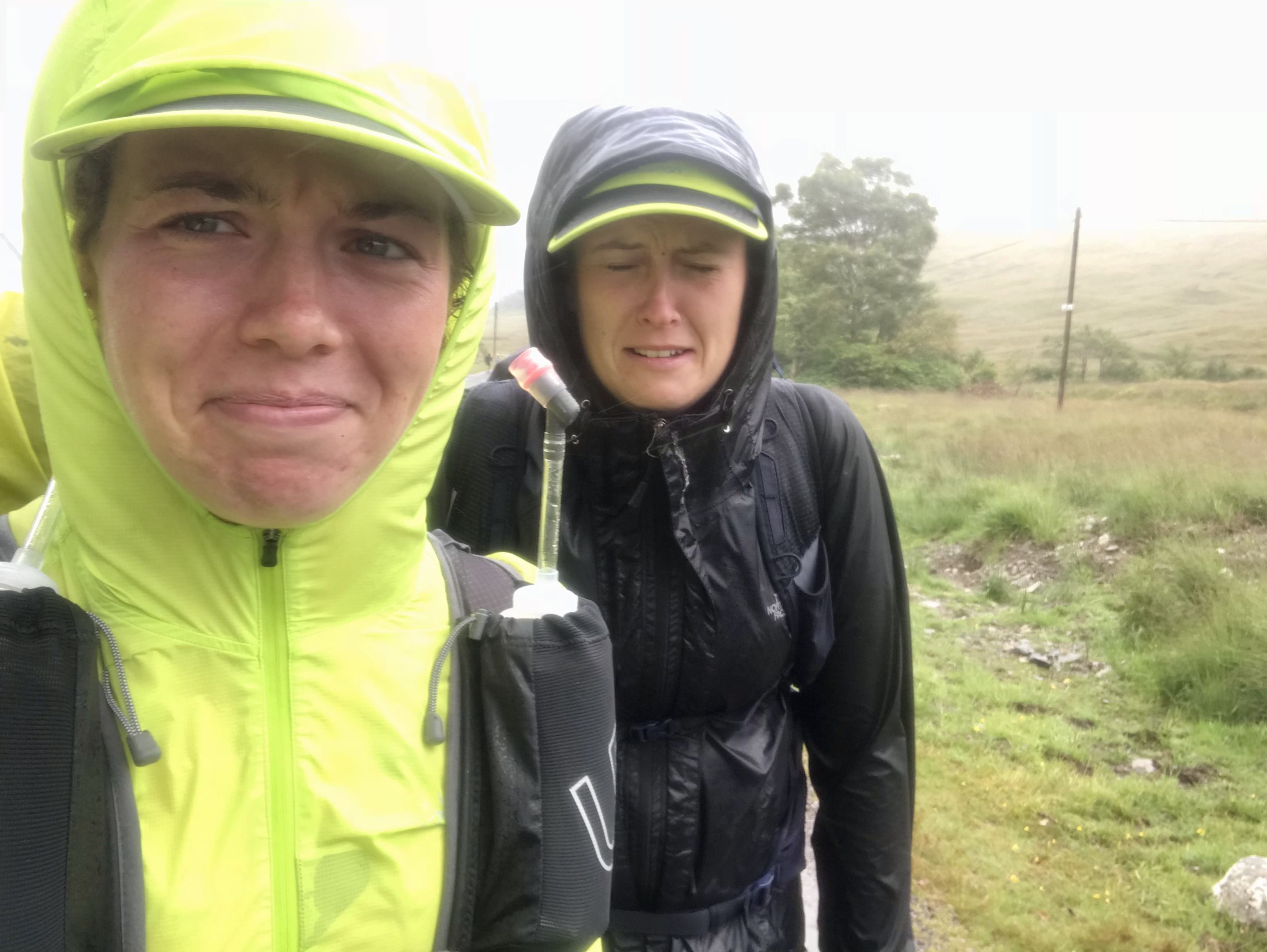 A selfie of Elise in a neon yellow jacket with the hood up with her friend also in hiking gear and hood up, both wincing at the rainy conditions