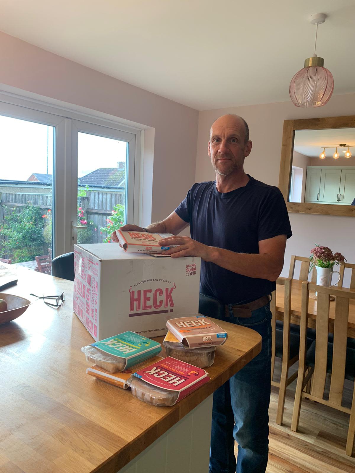 Elise's Dad Dave wearing a navy top and jeans, with a box full of packs of Heck sausages