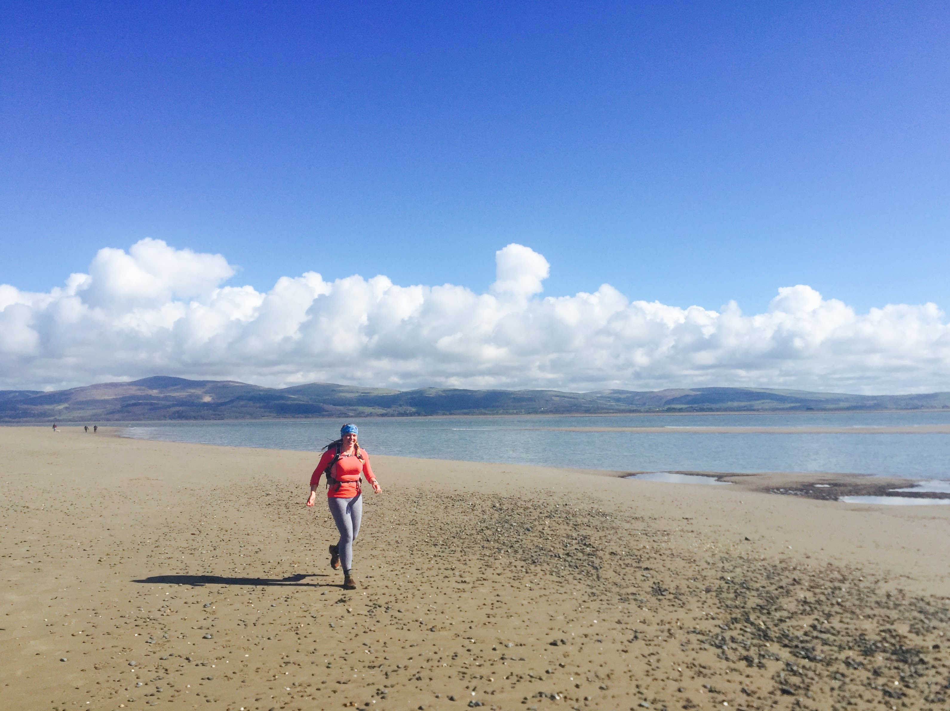 Elise Downing on the Wales Coast Path running on the beach in an orange top with a backpack underneath bright blue skies.