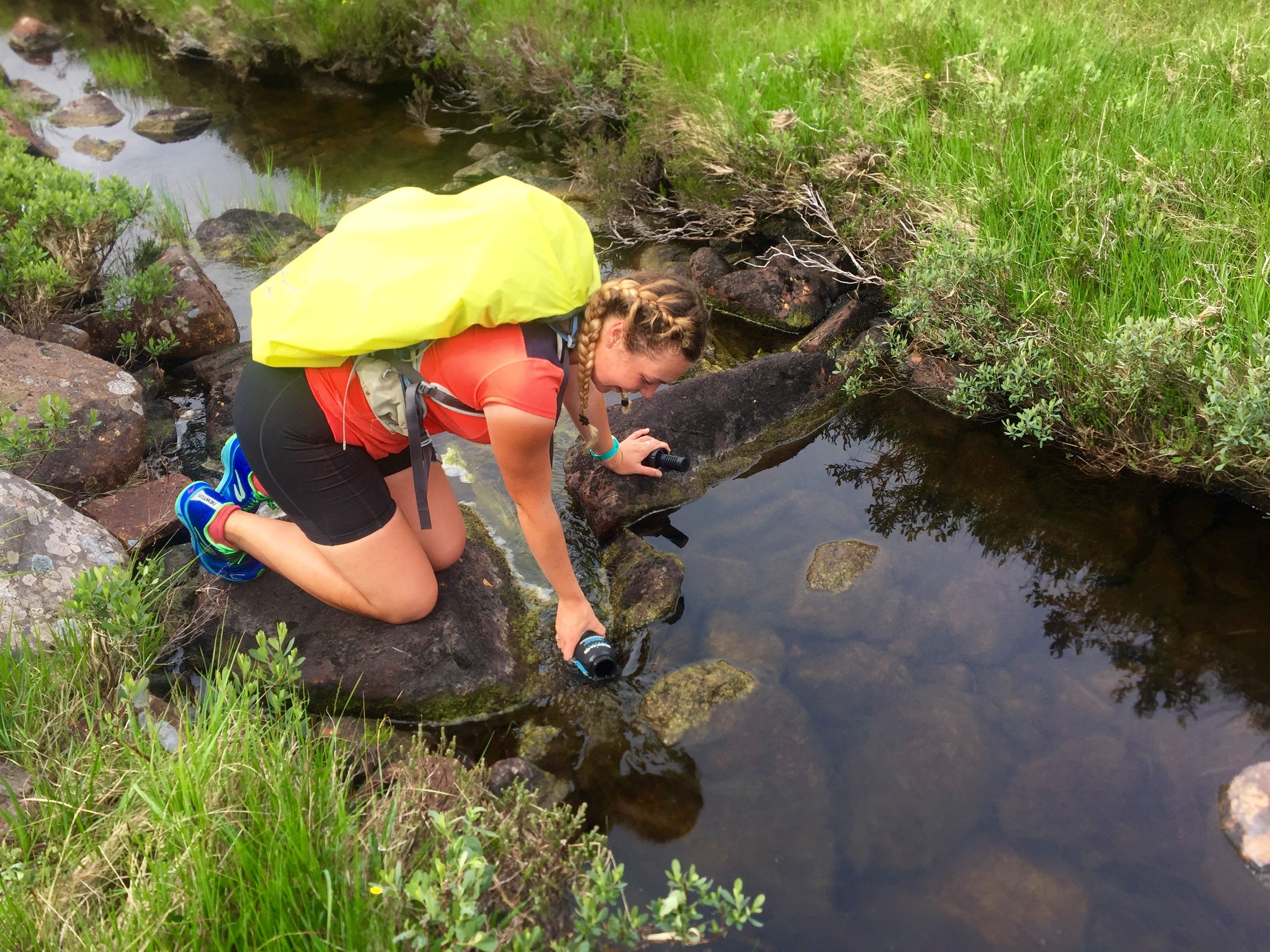 Elise wearing a backpack with a yellow cover filling her water up from a small river