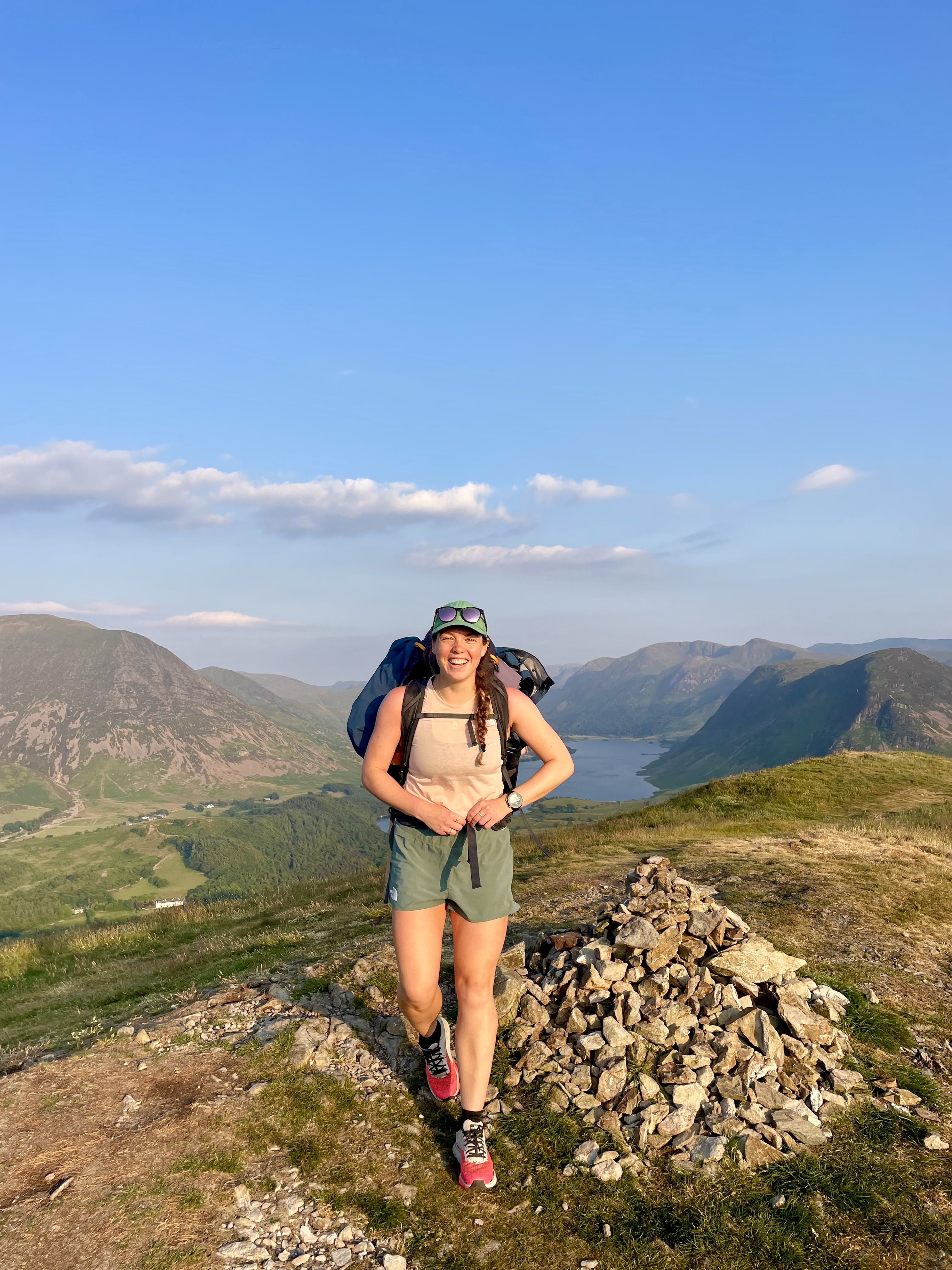 Elise in a light orange top, green shorts, green cap, and red and grey hiking boots,  smiling at the top of a mountain