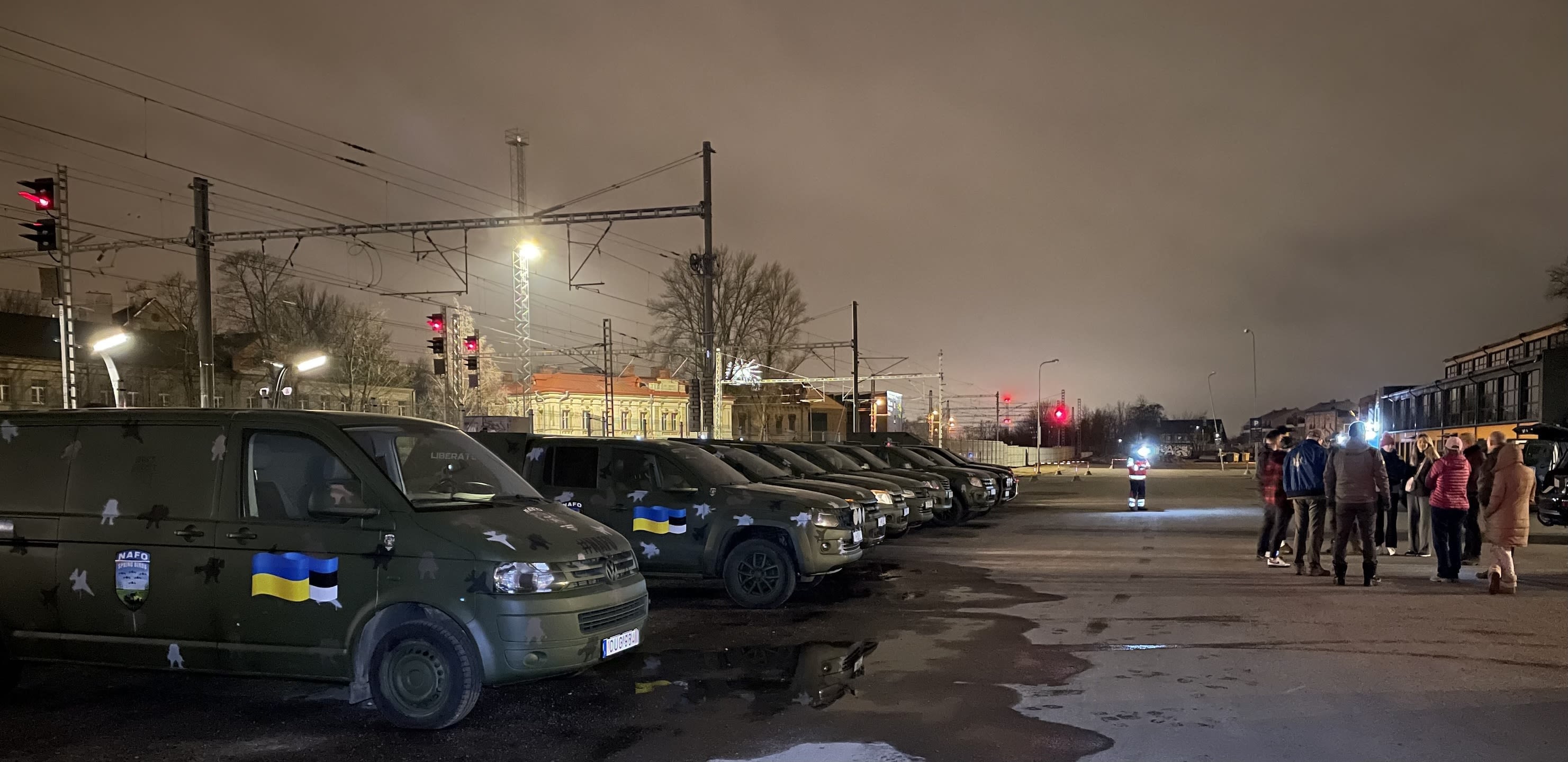 A line of vehicles painted military green in a parking lot on the left of the photo. A group of people huddled on the right-hand side. It is the early hours of the morning.